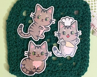 Könnte beinhalten: Drei graue Katzenaufkleber mit Glitzerakzenten auf einer grünen gehäkelten Oberfläche. Eine Katze trägt eine Kochmütze und hält eine Karotte.