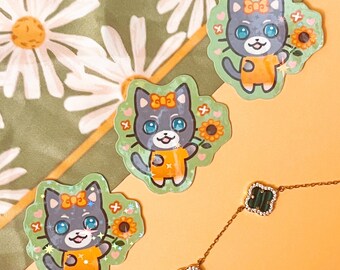 Könnte beinhalten: Drei Katzenaufkleber mit blauen Augen, orangefarbenen Hemden und Schleifen, die Sonnenblumen halten, sind auf einem grün-weiß gemusterten Gänseblümchen-Hintergrund angeordnet. Eine goldene Halskette mit grünen Kleeblatt-Anhängern befindet sich im Vordergrund.