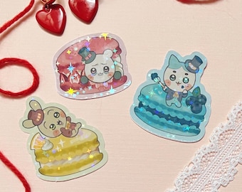 Könnte beinhalten: Eine Sammlung von Aufklebern mit niedlichen Cartoon-Charakteren auf Desserts, darunter ein Bär auf einem rosa Macaron, eine Katze auf einem blauen Kuchen und ein Hase auf einem gelben Kuchen. Außerdem ein Paar goldene Creolen mit roten herzförmigen Anhängern.
