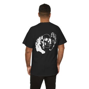 May include: Black t-shirt featuring a white graphic design of a skull and classical busts. The design is a split image, with the skull in the center. The t-shirt is made of a soft, comfortable material.