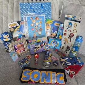 May include: A collection of Sonic the Hedgehog merchandise including a black t-shirt with large yellow text, a blue gift bag, a Lego set, a water bottle, stickers, a framed illustration, a plastic cup, a small toy, and various character-themed accessories.