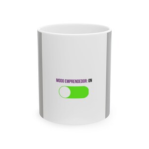 Puede incluir: Taza de cerámica blanca con borde y asa grises. La taza presenta el texto "MODO EMPRENDEDOR: ON" en morado, junto con un gráfico de interruptor de encendido/apagado verde. La taza está diseñada para bebidas calientes.