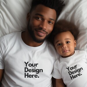 May include: A father and son wearing white t-shirts with the text "Your Design Here." printed on them. They are lying on a white bed.