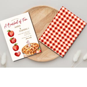 K&ouml;nnte beinhalten: Ein roter und wei&szlig;er karierter Hintergrund mit einer wei&szlig;en Einladungskarte darauf. Die Einladungskarte zeigt einen roten Apfelkuchen mit einer brennenden Kerze oben und einen roten Apfel mit Herzform unten. Der Text auf der Karte lautet "A Bushel of Fun Aspen is Turning 1".