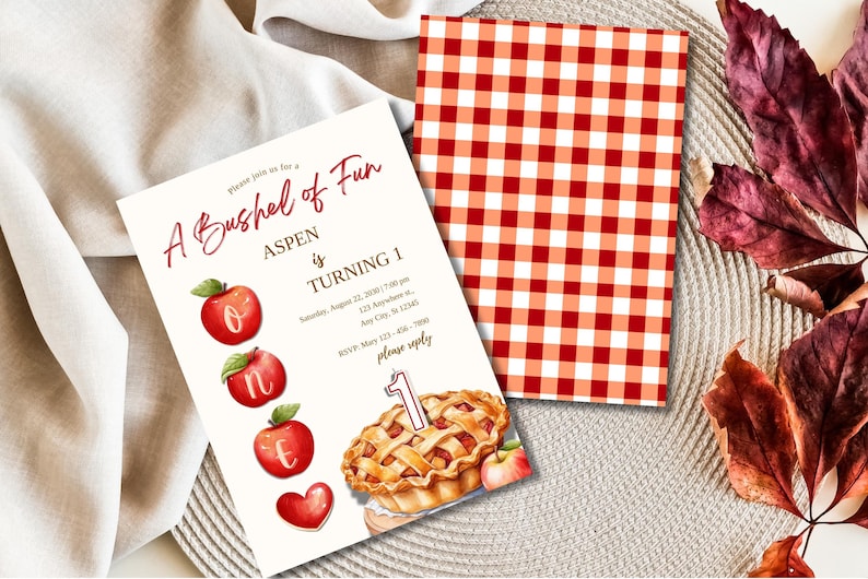 K&ouml;nnte beinhalten: Eine wei&szlig;e Einladungskarte mit roten &Auml;pfeln und einem Kuchen. Der Text lautet "A Bushel of Fun Aspen is Turning 1" mit Details f&uuml;r eine Party am 22. August 2020 um 19:00 Uhr. RSVP-Informationen sind enthalten.