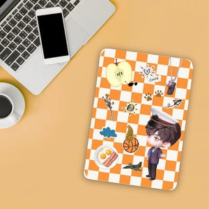 Puede incluir: Una funda para tableta a cuadros naranja y blanco con varias pegatinas, incluyendo una manzana, gafas de sol, un sol y una figura de dibujos animados con uniforme. También se ven un portátil, un teléfono y una taza de café.