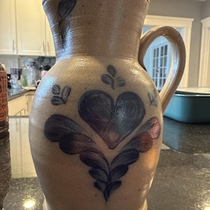 May include: A tan stoneware pitcher featuring a hand-painted blue heart motif with decorative leaf-like accents. The vessel has a wide, rounded body, a flared rim, and a sturdy side handle, showcasing a textured, earthy finish throughout the exterior.