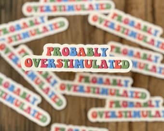 May include: A collection of stickers with the phrase "PROBABLY OVERSTIMULATED" in a retro font. The text is in red, pink, green, and blue, with a white border. The stickers are scattered on a wooden surface.