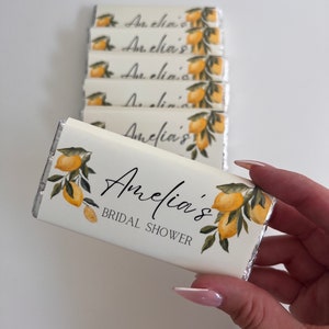 May include: A stack of white chocolate bar wrappers with a lemon and leaf design. The top wrapper says "Amelia's Bridal Shower".