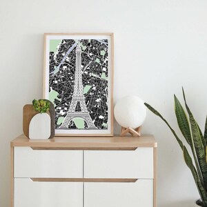 Peut inclure: Une carte de Paris en noir et blanc encadrée avec la Tour Eiffel au centre. La carte est sur une commode en bois clair avec une lampe de lune blanche et une petite plante verte.