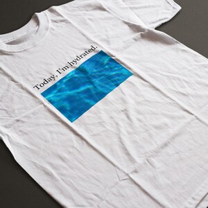 May include: White t-shirt with the text "Today, I'm hydrated" above a rectangular graphic of blue water. The shirt is made of a soft material and is displayed on a dark gray surface.