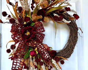 May include: A fall-themed wreath with a grapevine base, adorned with burgundy, gold, and cream-colored faux flowers, leaves, and berries. A large burgundy bow and a central burgundy peony add to the wreath's decorative appeal. The wreath is designed for door or wall decor.