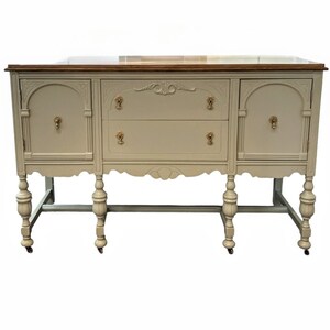 Peut inclure: Un buffet vintage de couleur blanc cassé avec deux portes et deux tiroirs. Le buffet a un dessus en bois et des pieds tournés avec des roulettes.