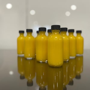 May include: A group of small, clear glass bottles with black lids filled with a bright yellow liquid. The bottles are arranged in a row on a reflective surface.