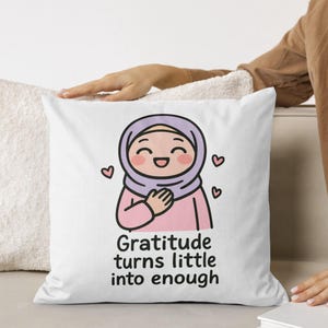May include: White square pillow with a cartoon illustration of a smiling person wearing a hijab and a pink outfit. The text "Gratitude turns little into enough" is below the illustration. Pink hearts are around the illustration.