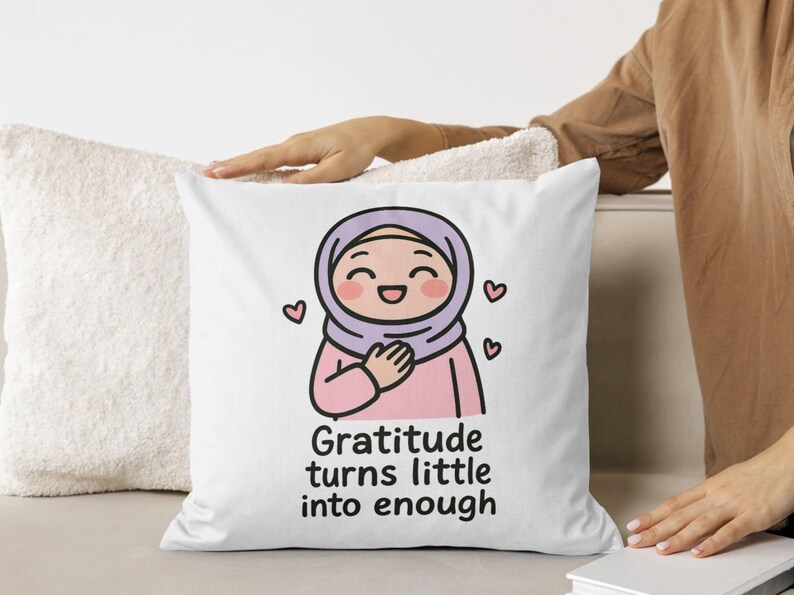 May include: White square pillow with a cartoon illustration of a smiling person wearing a hijab and a pink outfit. The text "Gratitude turns little into enough" is below the illustration. Pink hearts are around the illustration.