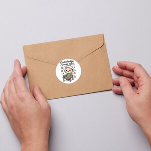 May include: A round sticker with a cartoon illustration of a person at a desk, with the text "Knowledge brings light." The sticker is on a brown envelope, held by two hands.