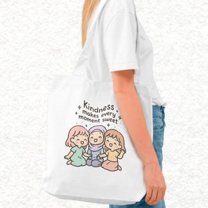 May include: White tote bag featuring an illustration of three cartoon girls and the text "Kindness makes every moment sweet." The girls are in pastel colors. The bag is carried by a person wearing a white shirt and blue jeans.