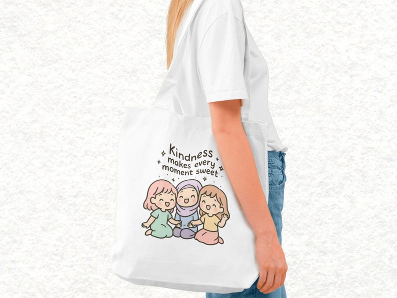 May include: White tote bag featuring an illustration of three cartoon girls and the text "Kindness makes every moment sweet." The girls are in pastel colors. The bag is carried by a person wearing a white shirt and blue jeans.