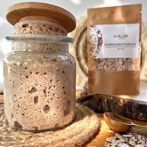 Pode incluir: Um frasco de vidro com tampa de madeira, cheio de um fermento de massa madre borbulhante castanho claro. Um pacote de papel castanho com a etiqueta "Solar Sourdough Starter Rye" está atrás do frasco. Uma colher de madeira e grãos também são visíveis.
