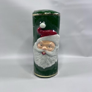 Puede incluir: Vela de pilar verde con una cara de Papá Noel en la parte delantera. La vela tiene un borde dorado alrededor de la parte superior e inferior.