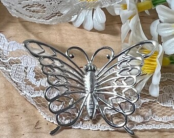 May include: A silver filigree butterfly brooch with delicate wings and a detailed body. The brooch is resting on a white lace fabric background.