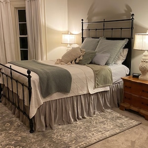 May include: A bedroom with a black metal bed frame, a white and grey patterned quilt, and a grey bed skirt. The bed has grey and green pillows. Two lamps with white bases and beige shades sit on a wooden bedside table.
