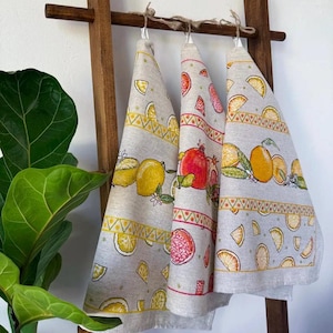 May include: Three linen kitchen towels with fruit designs hang on a wooden rack. The towels feature lemons, pomegranates, and oranges in vibrant colors. Each towel has a decorative border with geometric patterns.