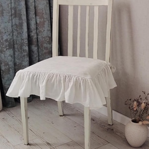 May include: A white wooden chair with a slatted back and a square seat. The seat is covered with a white, ruffled fabric cover. The chair has a distressed, vintage look, suitable for farmhouse or shabby chic decor.