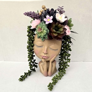 May include: A tan-colored head-shaped planter featuring a face with closed eyes and hands resting under the chin. The planter is filled with an arrangement of artificial succulents, small pink and white flowers, and long trailing green vines.