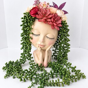 Può includere: Un vaso a forma di testa color beige con occhi chiusi, ciglia lunghe e mani sotto il mento. La parte superiore è decorata con un bouquet di fiori artificiali rossi, rosa e crema, con lunghi rami verdi ricadenti di piante grasse.
