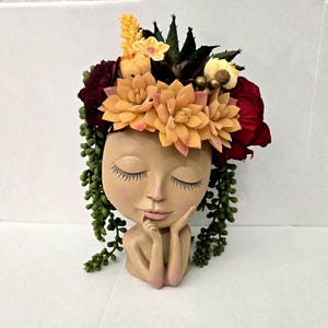 May include: A tan-colored head-shaped planter featuring a serene face with closed eyes and long eyelashes. The figure rests its chin on its hands. It is adorned with a vibrant arrangement of artificial peach succulents, deep red flowers, and green vines.