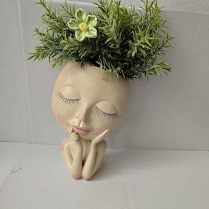 Puede incluir: Una maceta de cerámica beige con forma de cabeza que muestra un rostro sereno con ojos cerrados, pestañas largas y mejillas rosadas. La figura apoya la barbilla sobre sus manos. Una planta artificial verde con una flor clara está colocada dentro.
