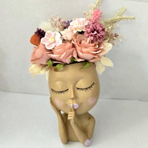May include: A tan-coloured head-shaped planter featuring a face with closed eyes and a finger held to the lips in a shushing gesture. The planter is filled with an arrangement of artificial flowers in shades of pink, cream, and purple.