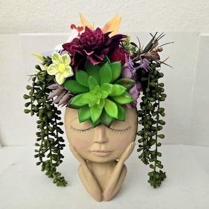 May include: A beige human-head shaped planter with closed eyes and hands resting on the chin. The planter is filled with an arrangement of artificial green succulents, a large dark red dahlia, a small yellow flower, and trailing green vines.