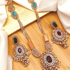 Puede incluir: Un conjunto de collar y pendientes de oro con piedras multicolores y detalles de perlas. El collar tiene una cadena larga con múltiples hebras y un colgante con una piedra roja grande rodeada de piedras más pequeñas. Los pendientes tienen un diseño similar con una piedra roja más pequeña y gotas de perlas.