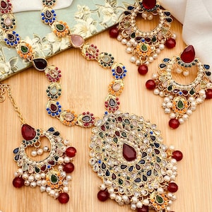 May include: A gold-tone Indian bridal jewelry set with red, green, blue, and white stones. The set includes a necklace, earrings, and a maang tikka.