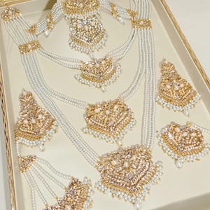 Puede incluir: Un conjunto de joyas de novia indias tradicionales con un diseño de oro y perlas blancas. El conjunto incluye un collar, pendientes y una diadema.
