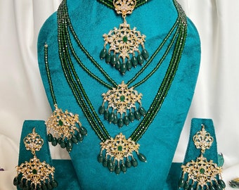 Puede incluir: Un conjunto de collar de cuentas verdes y doradas con múltiples hileras, un collar gargantilla y pendientes a juego. El collar presenta intrincados diseños florales y cuentas verdes colgantes. El conjunto se muestra en un soporte de terciopelo turquesa. "My Jewel Box" está escrito en una pequeña etiqueta.