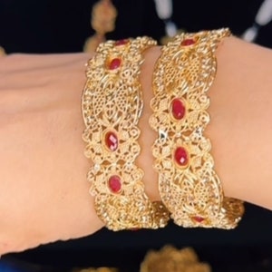 Puede incluir: Dos brazaletes de oro con intrincados diseños de filigrana y piedras preciosas rojas. Los brazaletes están apilados en la muñeca de una persona.