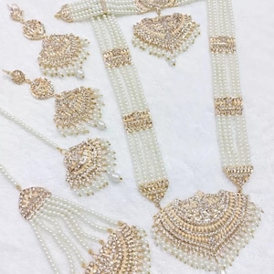 May include: A traditional Indian bridal jewelry set featuring a long pearl necklace with a gold pendant, a matching choker, earrings, and a maang tikka. The set is adorned with sparkling stones and intricate designs.