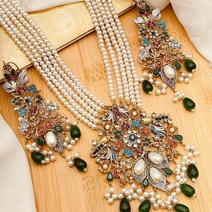 May include: A multi-strand pearl necklace with a large, ornate pendant featuring a floral design with colorful stones and green accents. The pendant is attached to a chain with a pearl drop. The necklace is paired with matching earrings with a similar design.