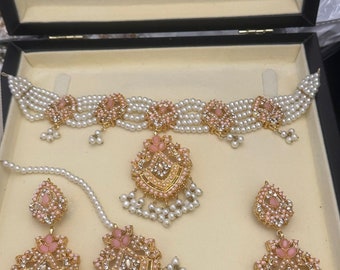 May include: An elegant jewelry set featuring a choker necklace, earrings, and a headpiece, all presented in a black box. The set includes faux pearls, gold-toned metal, and pink accents. The earrings and headpiece have dangling pearl strands.