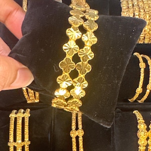 Puede incluir: Brazaletes dorados con diseño floral, exhibidos sobre una superficie negra. Los brazaletes presentan un patrón repetitivo de flores de cuatro pétalos. Otros brazaletes dorados son visibles en el fondo.