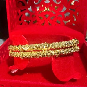May include: Two gold bangles with intricate floral and scrollwork designs are displayed inside a red velvet jewelry box. The bangles are stacked on top of each other, showcasing their detailed craftsmanship. The box is open, revealing a heart-shaped cutout design.