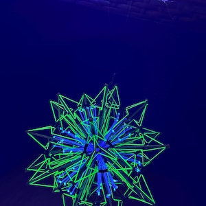 May include: A geometric, star-shaped light fixture with a complex design. The fixture features a network of green, triangular shapes and blue lights, set against a dark blue background. The overall effect is a modern, artistic display.