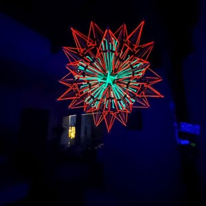 May include: A geometric, star-shaped light fixture with a complex, multi-layered design. The outer layer is outlined in bright red, while the interior features green and blue accents. The fixture is suspended from the ceiling in a dark room.