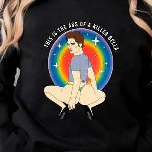 Peut inclure: Sweat-shirt noir avec un dessin color&eacute; d'une personne accroupie, entour&eacute;e d'un cercle arc-en-ciel et du texte "THIS IS THE ASS OF A KILLER BELLA". L'illustration est de style cartoon.