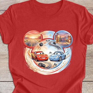 Peut inclure: T-shirt rouge avec un motif de dessin anim&eacute; de deux voitures de course, une rouge et une bleue, sur une piste. Le motif comprend un coucher de soleil dans le d&eacute;sert et une sc&egrave;ne de ville, avec des traces de pneus et des drapeaux &agrave; damier. Une tasse de caf&eacute; est en bas &agrave; gauche.