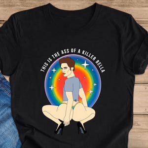 Peut inclure: T-shirt noir avec une illustration de dessin anim&eacute; d'une personne accroupie, portant une chemise bleue, des talons et des sous-v&ecirc;tements. Le motif est plac&eacute; sur un fond aux couleurs de l'arc-en-ciel avec des &eacute;toiles et le texte "THIS IS THE ASS OF A KILLER BELLA" en cercle.